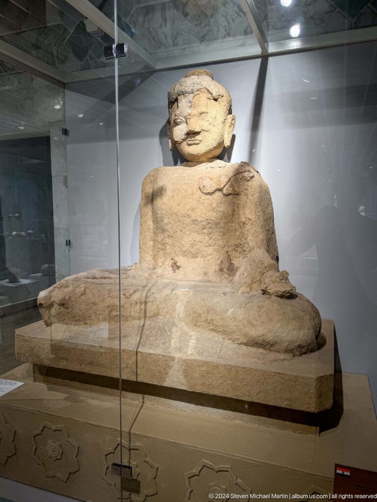 Seated pale stone figure behind glass.