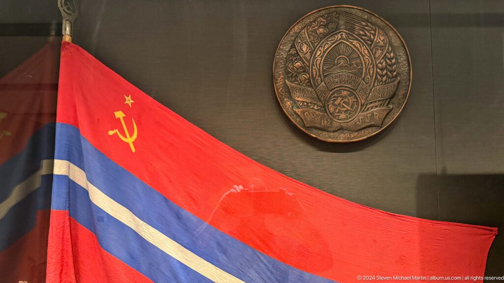 Red and blue Soviet Kyrgyz flag beneath round emblem.