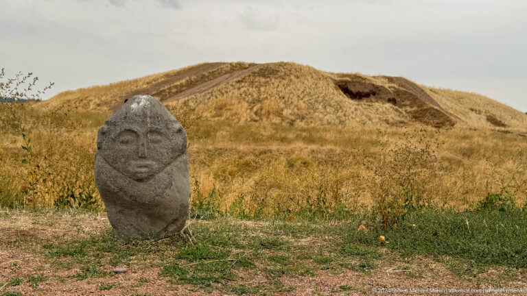 A carved stone figure stands in dry grass before a low mound associated with ancient Balasagun