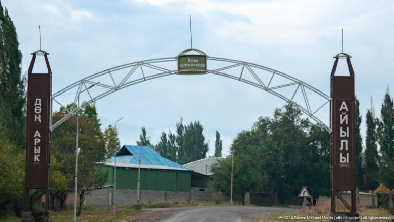 A metal arch with Cyrillic lettering marks the entrance to a tree-lined settlement