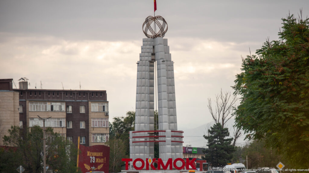A tall pale monument rises above large red Cyrillic lettering in a city streetscape