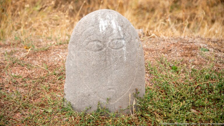 A pale rounded memorial stone bears a simplified human face and shallow carved marks