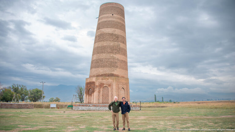 Two visitors stand side by side in the open field with the minaret behind them