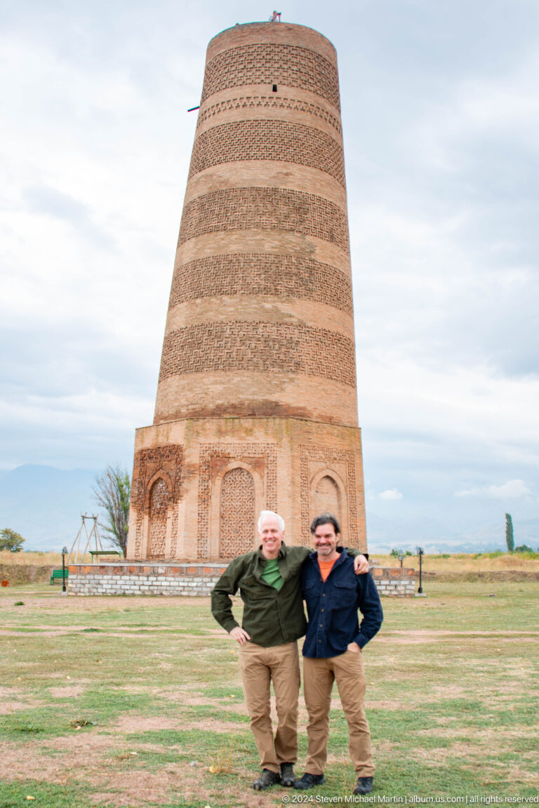 Two visitors smile in a close portrait with the brick minaret rising behind them