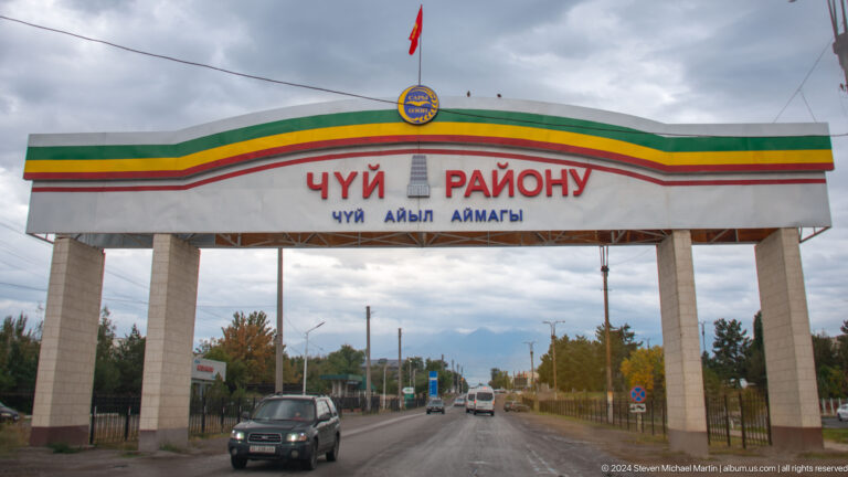 A broad roadside gateway spans the highway with Kyrgyz and Russian district lettering