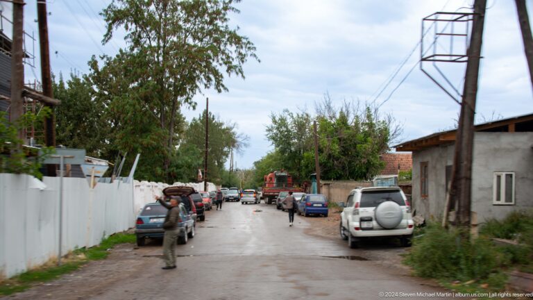 A quiet unpaved street is lined with low houses, utility poles, parked vehicles, and trees