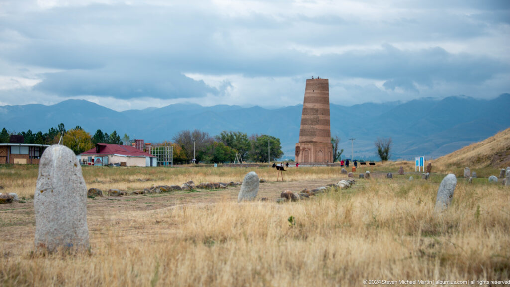 The minaret rises beyond scattered carved stones and dry grass