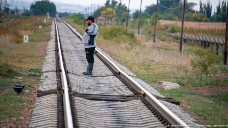 A person carefully steps across parallel railway rails in an open rural area