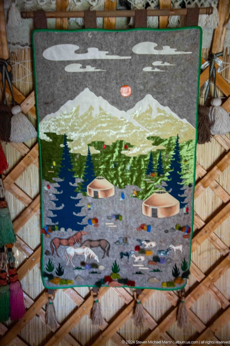 A textile depicts mountains, trees, water, and yurts in a stylized landscape
