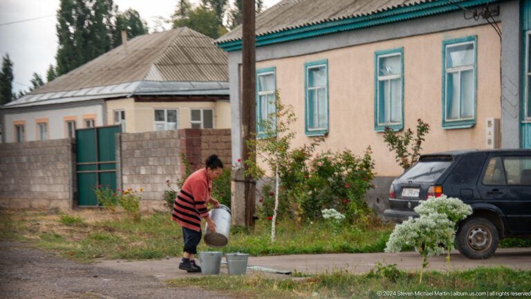 A woman walks beside a modest blue-trimmed house while carrying a bucket