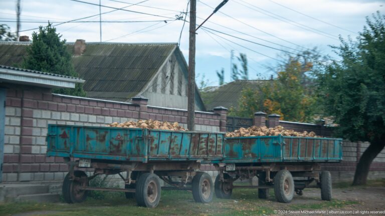 Two long farm trailers stand beside a low rural dwelling