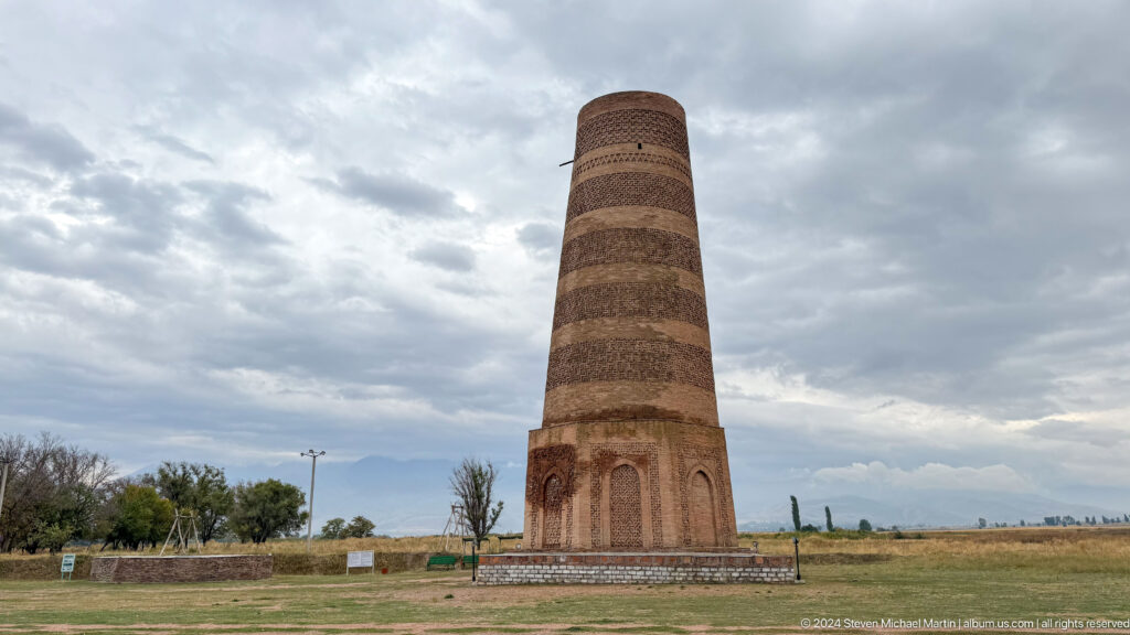 The minaret stands alone in the grassland with mountains beneath a cloudy sky