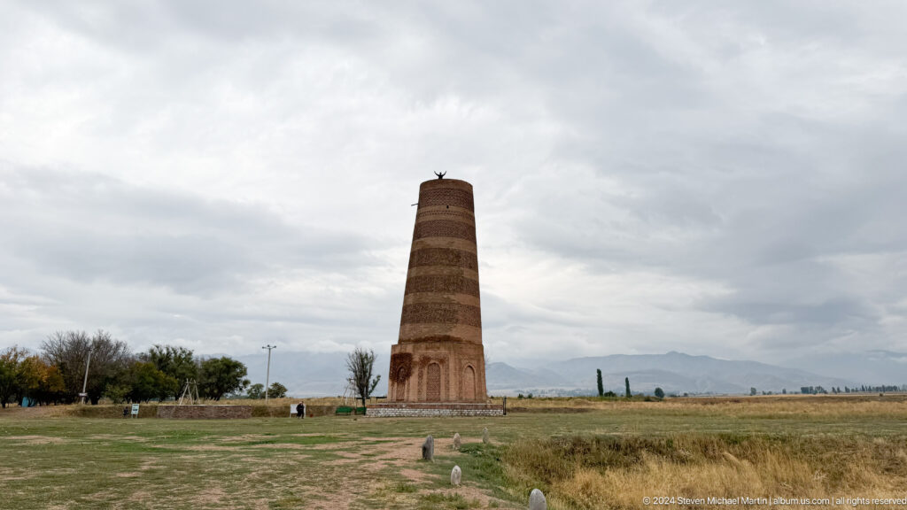 The minaret stands far across the grassland under a broad cloudy sky