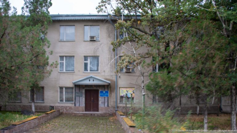Plain three-story building partly hidden by trees.