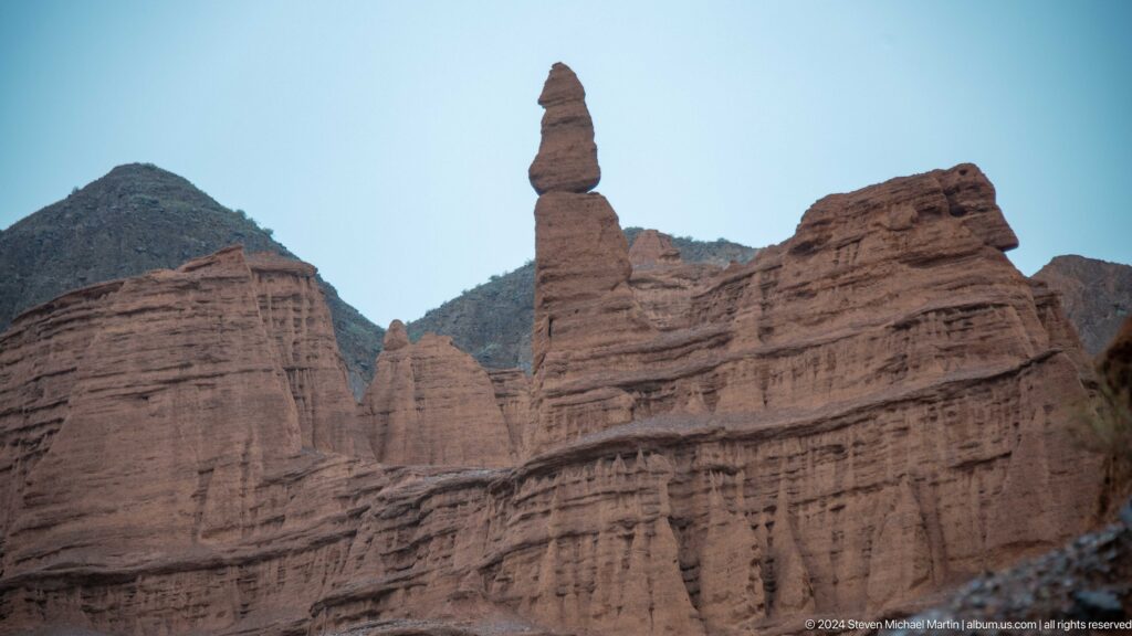 Tall slender pinnacle rising above banded red cliffs.