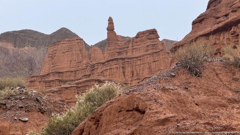 Tall red pinnacle and banded cliffs under blue sky.