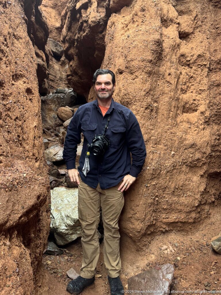 Bearded man standing between canyon walls with camera gear.