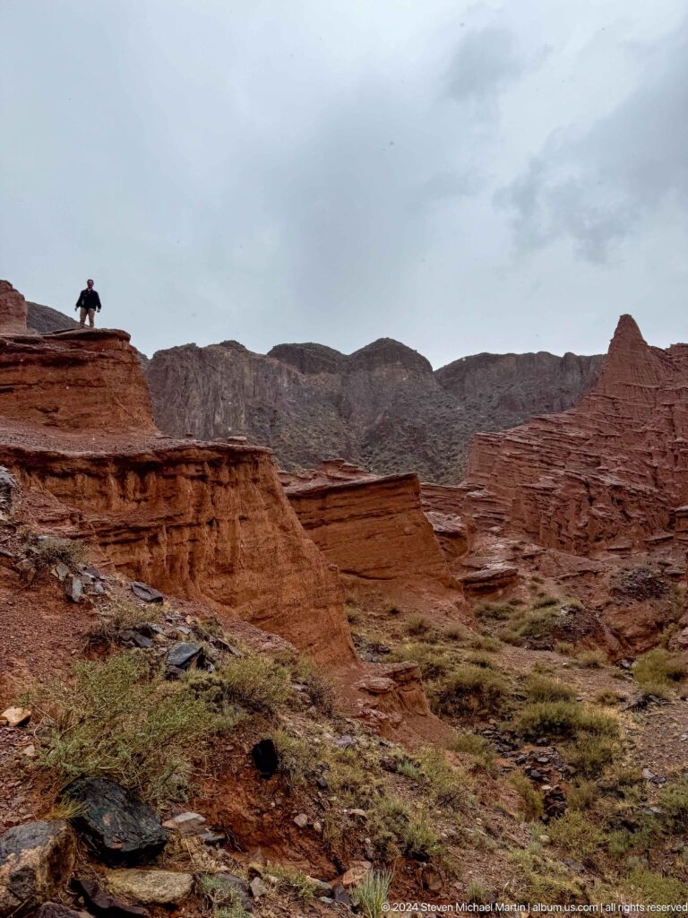 Wide red ridge and isolated pointed formation beneath gray mountains.