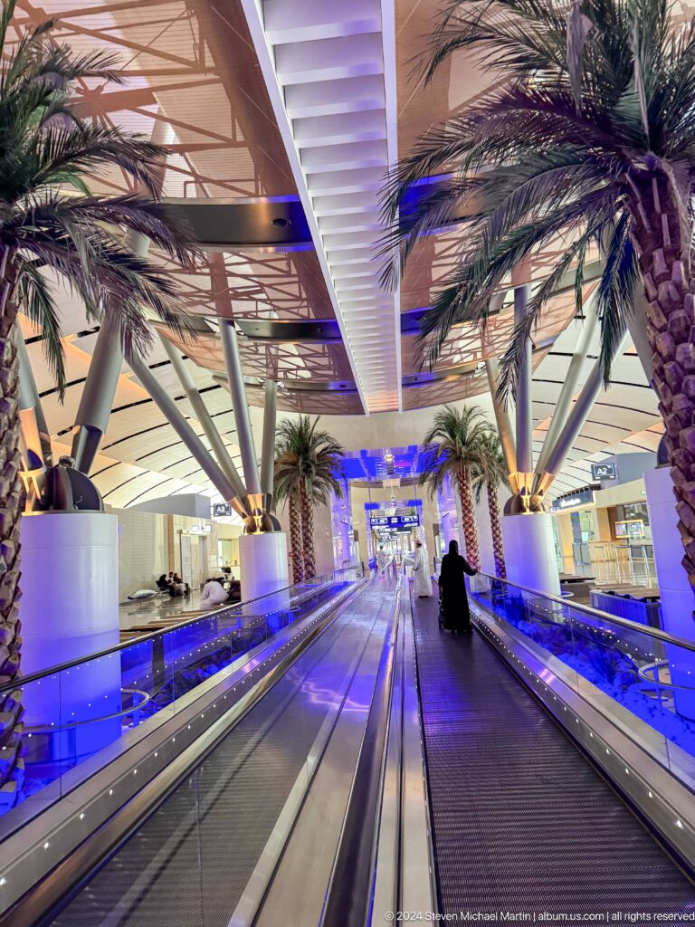 Blue-lit airport concourse with palm trees and moving walkway in Muscat, Oman.