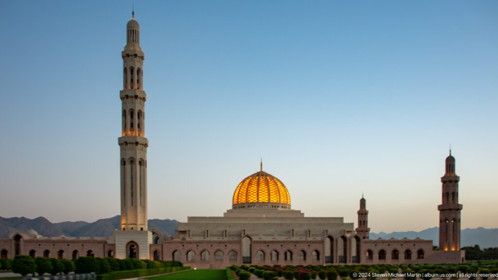 Wide mosque facade with dome and minarets beneath blue sky in Muscat, Oman.