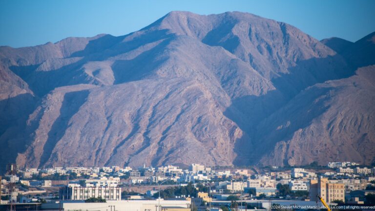 Dense white city beneath steep sunlit mountain in Muscat, Oman.