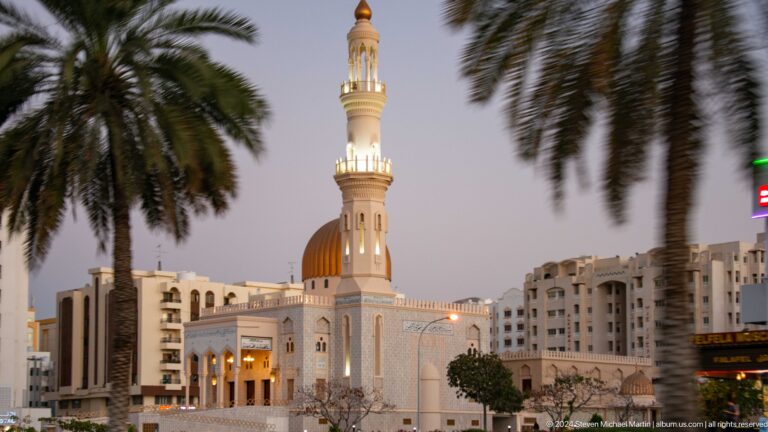 White mosque with gold dome framed by palms and city buildings in Muscat, Oman.