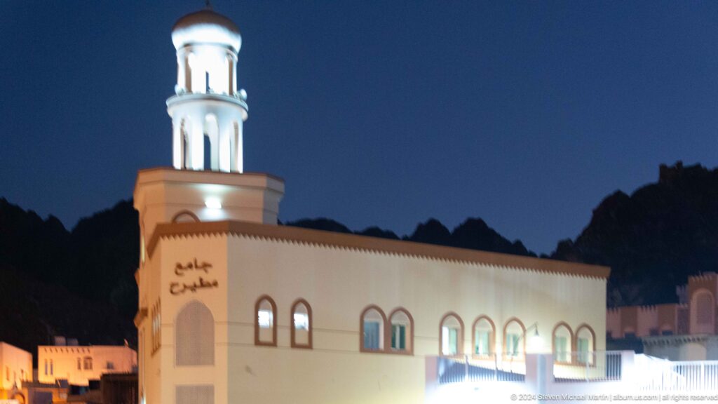 White mosque with illuminated minaret against mountains.