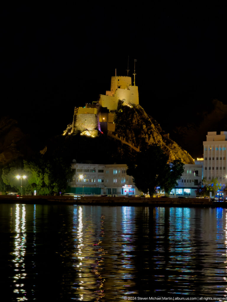 Mutrah fort glowing above waterfront at night.