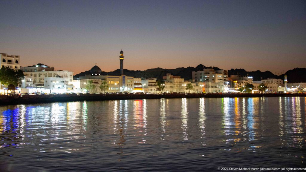 Wide mutrah waterfront skyline reflected in water.