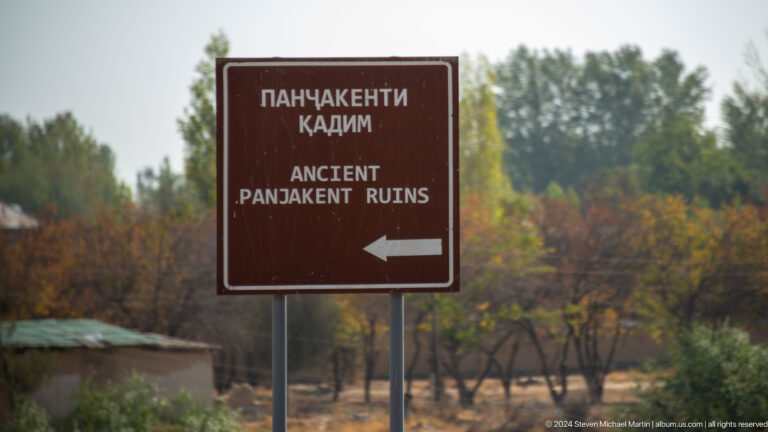 Bilingual brown road sign with a left-pointing arrow indicating the entrance to the Ancient Panjakent Ruins in Tajikistan, with roadside vegetation in the background.