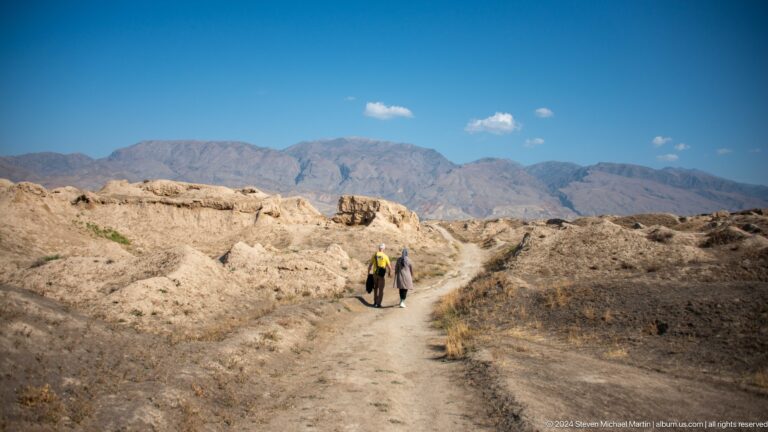 Two visitors walk a dirt path between low earthen ruins at Ancient Panjakent, with hills and distant mountains under a hazy sky.
