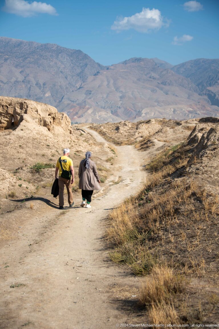 Rear view of two visitors walking along a narrow dirt path between steep, eroded earthen banks at the Ancient Panjakent ruins.