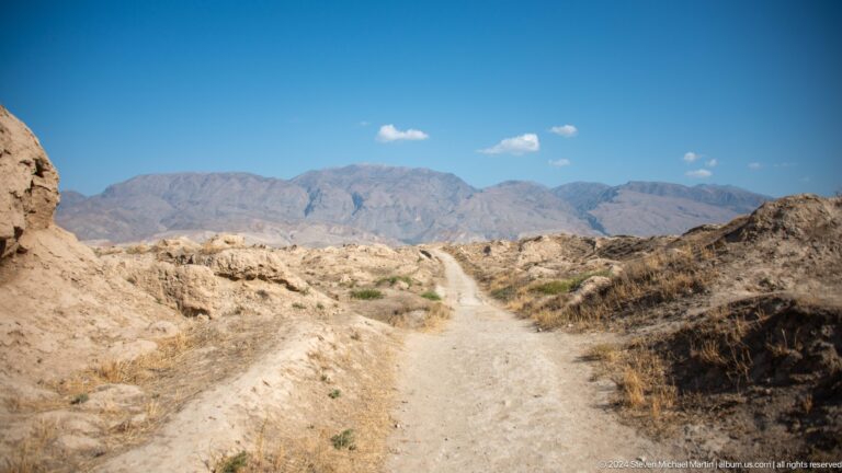 Landscape view of a winding archaeological path crossing low, eroded mounds at Ancient Panjakent with mountains in the background.