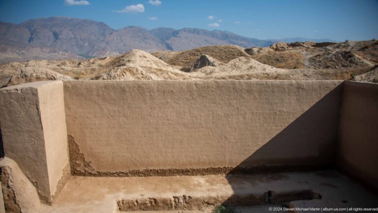 Close view of a conserved mud-brick wall corner with a smooth protective coating and a triangular shadow against rough excavated earth at Ancient Panjakent.