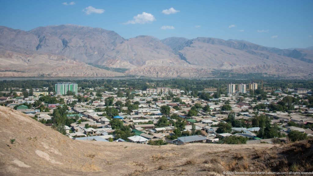 Panoramic view from an elevated archaeological hill looking over the modern city of Panjakent spread across a green valley with mountains in the distance.