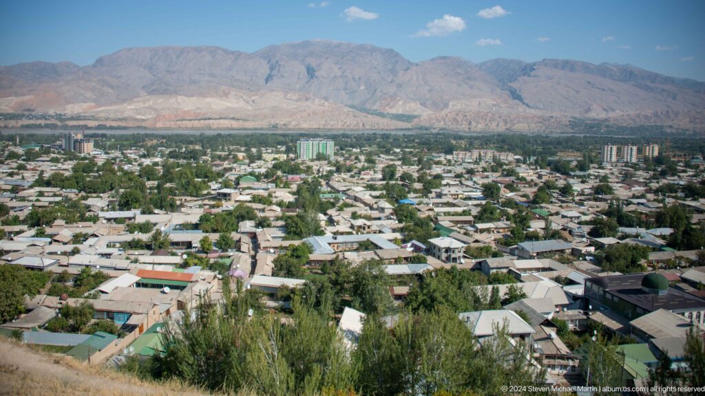 Elevated view over modern Panjakent with green fields on the valley floor and arid mountains beyond, seen from a dry archaeological hill.