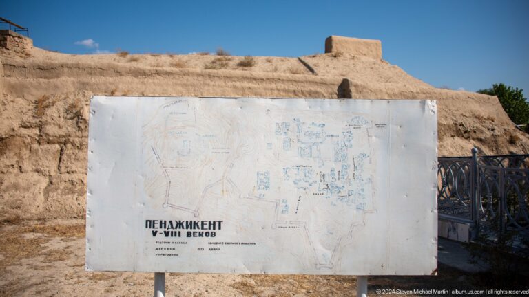 Weathered white information board with a faded site plan of Ancient Panjakent standing before sunlit earthen ruins; much of the map text is unreadable.