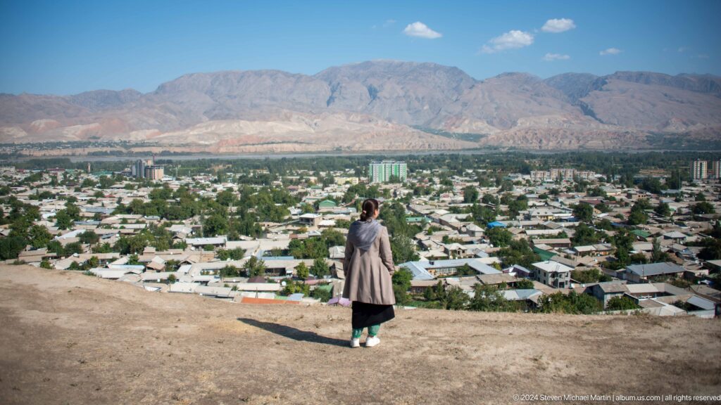 Person standing at an overlook on a hill above ruins, looking out over the modern city of Panjakent with mountains in the distance.