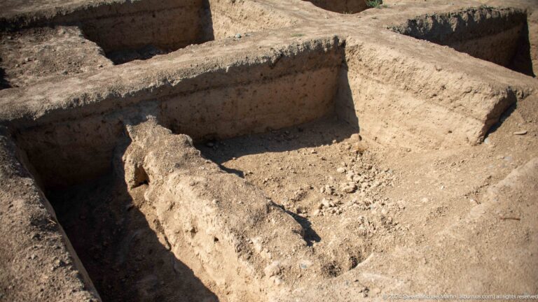 Top-down view into a rectangular archaeological cut with low mud-brick and earthen walls forming a partial interior space within the ruins.