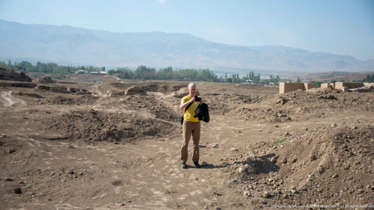 Single visitor standing among the low earthen ruins of Ancient Panjakent at an archaeological site in Tajikistan, with a distant valley beyond.