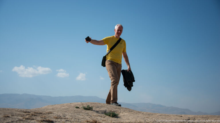 Low-angle view of a visitor with a raised camera on a ridge at Ancient Panjakent, silhouetted against blue sky and distant mountains.