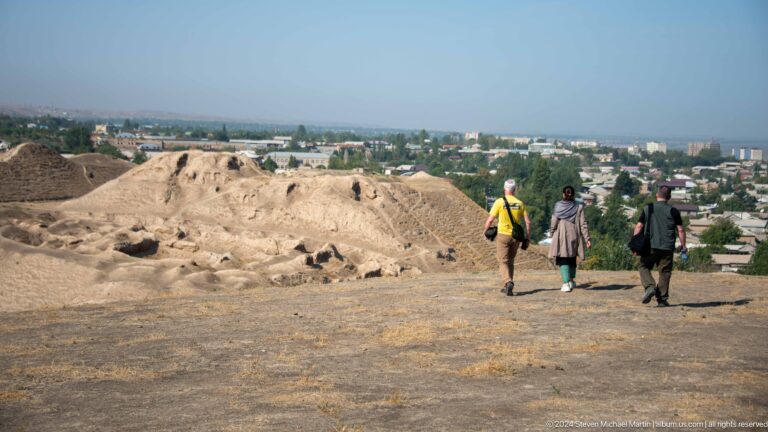 Three visitors walk along the earthen ridge atop the exposed hilltop ruins of Ancient Panjakent with the city visible in the distance.