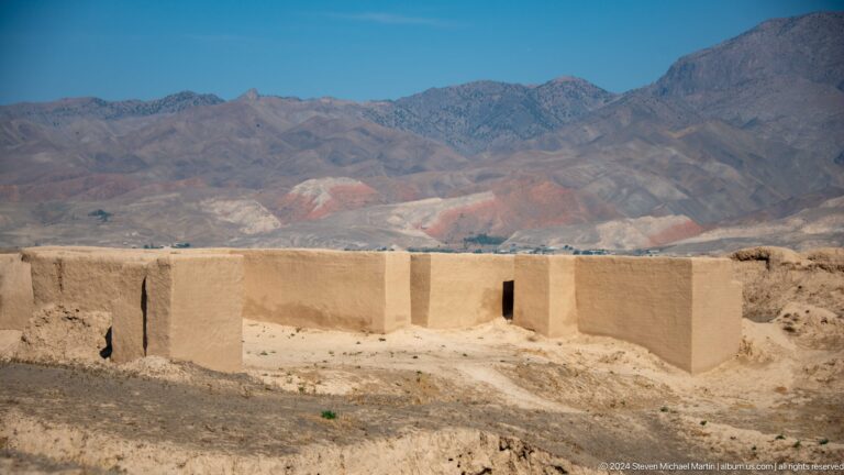 Wide view of reconstructed earthen walls outlining an ancient building footprint at Ancient Panjakent, with mountains in the background.