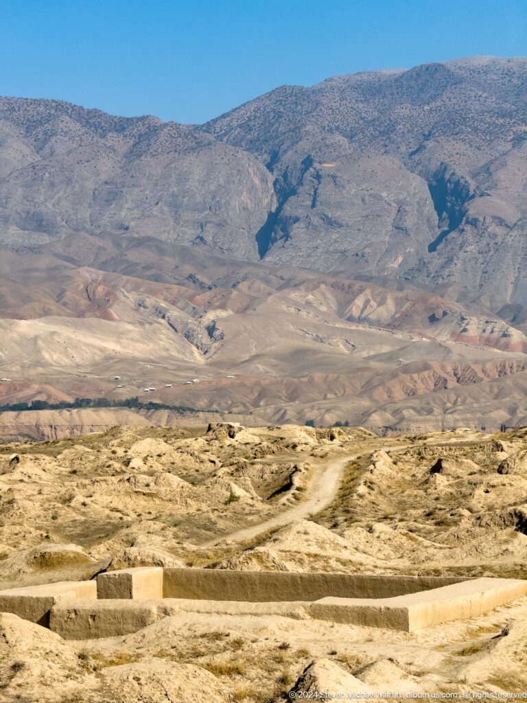 Telephoto view of a winding path crossing low archaeological mounds with folded mountains rising in the background.