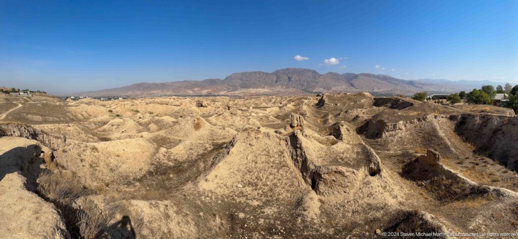 Wide view of an excavated and eroded sector of Ancient Panjakent, showing channels, low earthen walls, and exposed deposits against a broad mountain horizon.