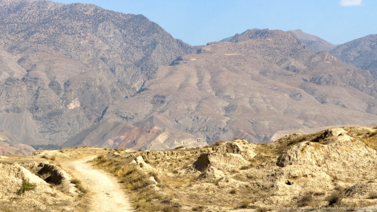 Curving path through the low archaeological mounds of Ancient Panjakent with steep mountains in the distance.
