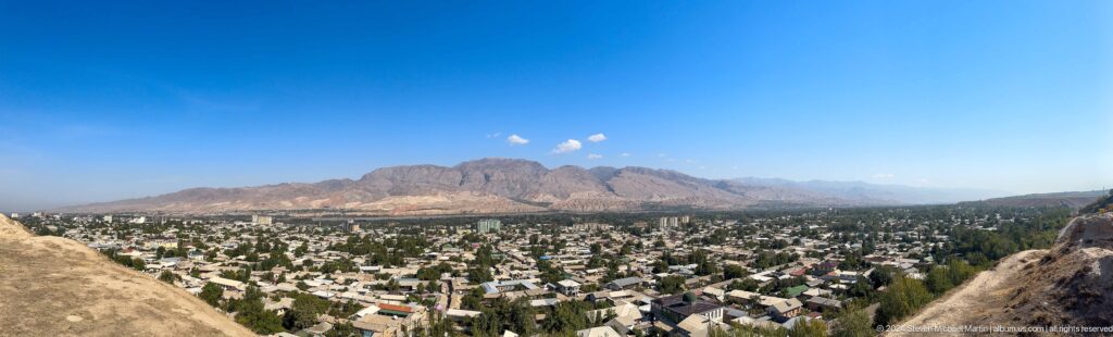 Panoramic view of modern Panjakent spread across a green valley, with distant mountain ridges rising behind the city, seen from an elevated archaeological hill.