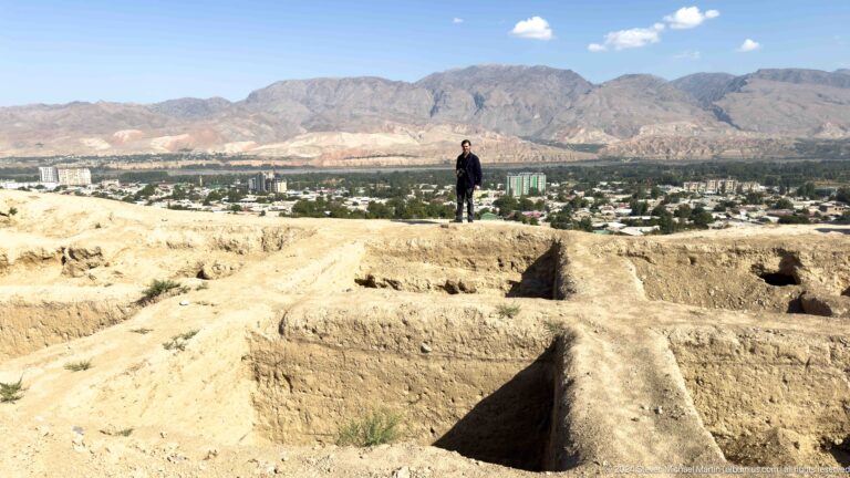 Visitor standing on the edge of Ancient Panjakent’s excavated rooms with the modern city of Panjakent visible in the background.