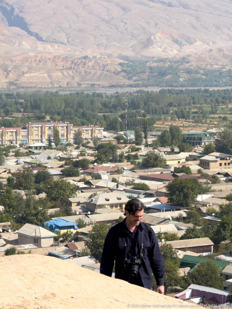 Partially visible person at a hilltop overlook with the dense, tree-filled cityscape of modern Panjakent in the background.