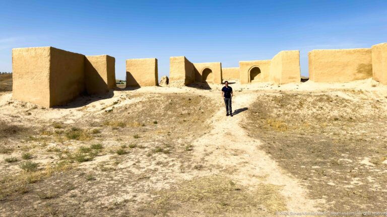 Wide view of a visitor approaching a conserved earthen-walled structure with multiple arched openings at Ancient Panjakent.